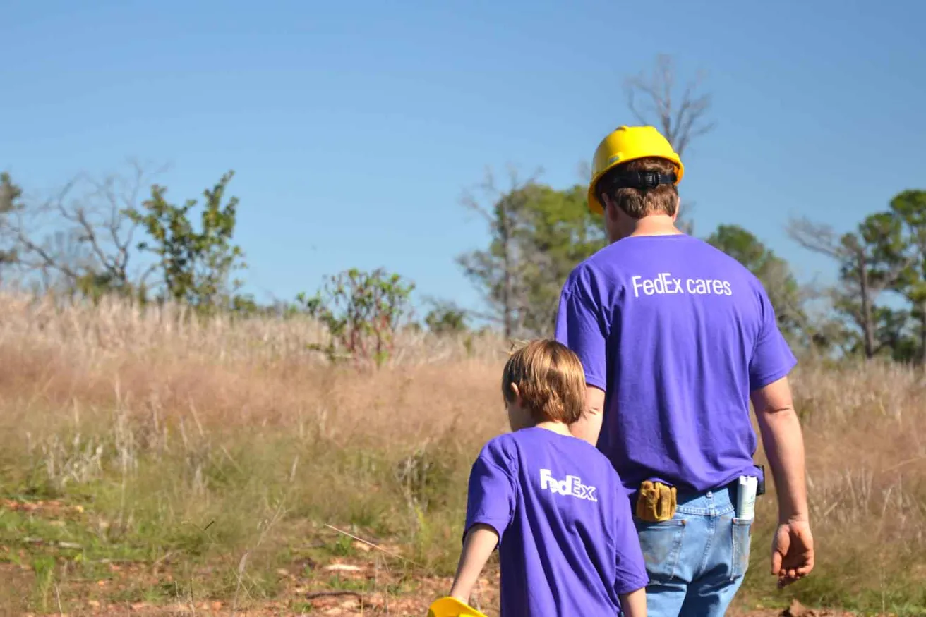 A man in yellow hard hat and blue FedEx t-shirt walks with a young boy in a forest.