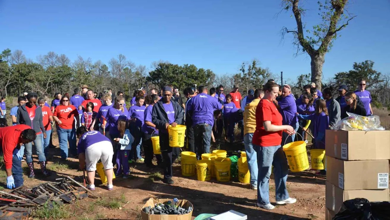 A group of people in purple shirts gathered around a stack of yellow buckets, getting ready to plant trees.