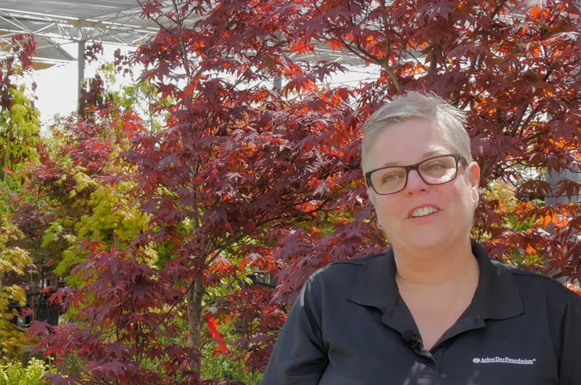 Woman speaking to a camera in front of colorful trees.