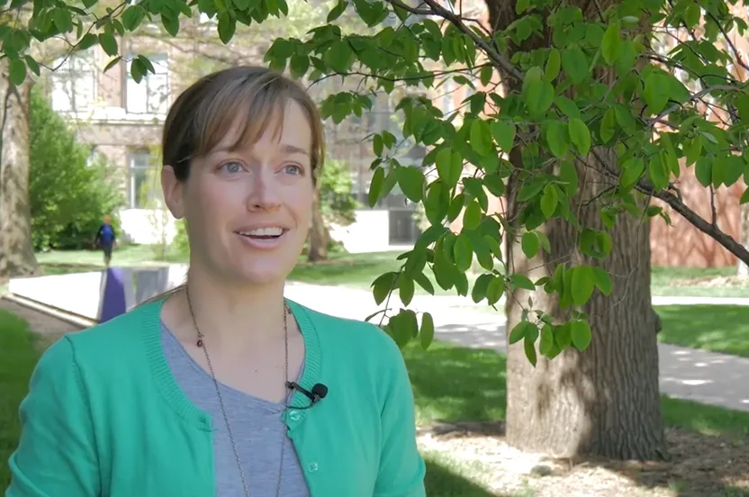 Woman speaking to a camera in front of a tree.