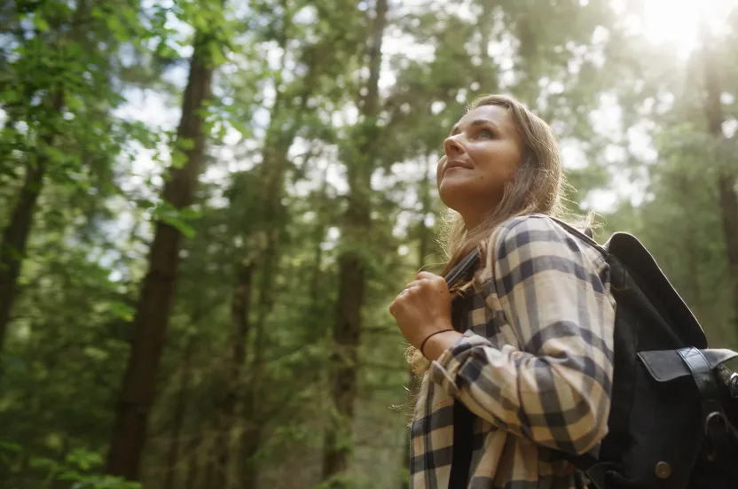 A woman hikes through the woods surrounded by trees and dappled sunlight, embodying a sense of hope amidst the natural beauty around her.